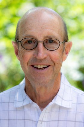 Headshot of Graham Hollis outdoors in front of soft green foliage, wearing glasses and a light shirt, smiling at the camera.