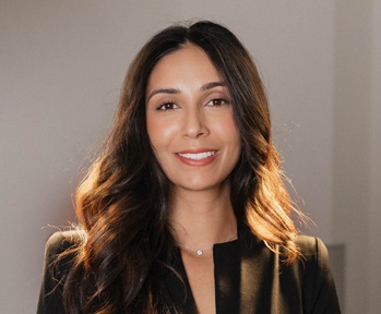 Close-up portrait of Dr. Leela Mundra smiling at the camera, wearing a black top and a delicate necklace. Soft light highlights her hair and face against a neutral indoor background.